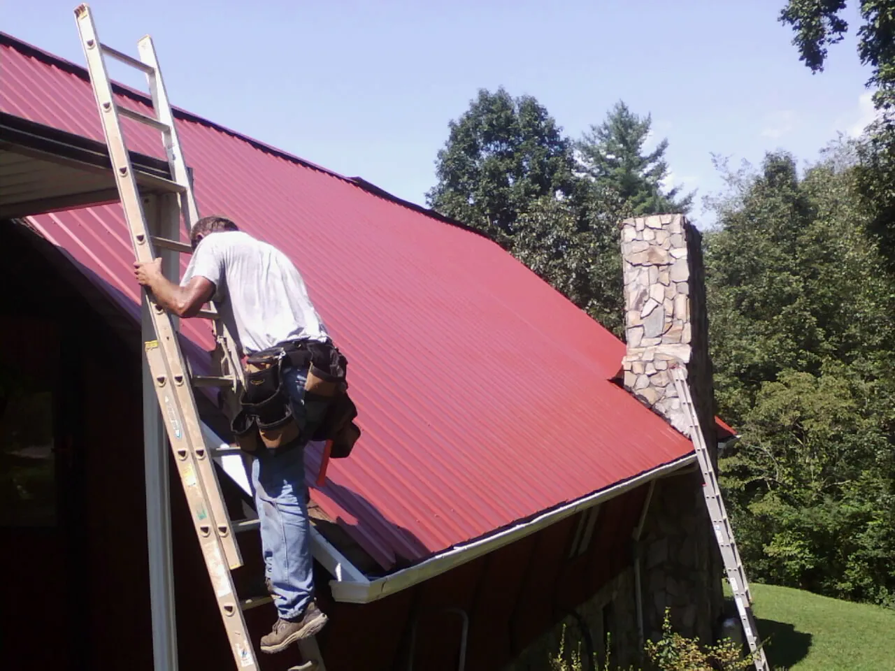 Roofer on a ladder beside a red metal roof and stone chimney — Soffit & Fascia Repair by Roofing Brothers in North East